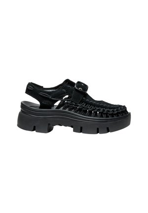 Sandale noire tressée avec semelle épaisse à crampons, bride réglable avec boucle et design à talon ouvert sur fond blanc.
