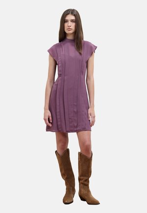 Giovane donna in piedi di fronte a noi che indossa un vestito viola senza maniche con cuciture verticali e stivali marroni in camoscio al ginocchio.