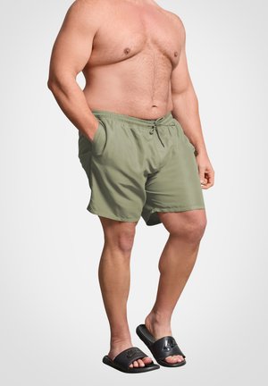 Homme portant un short de bain vert olive avec les mains dans les poches et des sandales Nike noires, debout devant un fond uni.