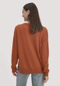 Rustfarbener Pullover aus weichem Stoff, mit lockerer Passform, gerippten Bündchen und einem leicht verlängerten Rückensaum. Getragen mit hellblauen Jeans.