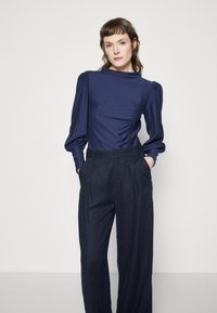 Navy blue blouse with puff sleeves; smooth texture and gathered detailing on the side. Paired with high-waisted, wide-leg navy trousers.