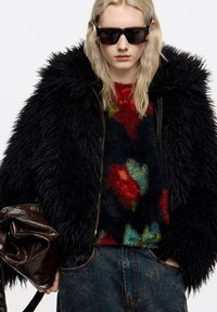 Czarna kurtka z sztucznego futra z wysokim kołnierzem, zestawiona z teksturowanym, kolorowym swetrem z abstrakcyjnymi wzorami oraz ciemnymi jeansami.