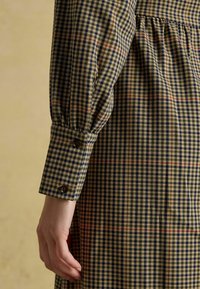 Ilgi piedurkņu pārbaudīta kleita ar bēšas, tumši zilas un sarkanas krāsas rakstu; ar pogām apakšā un savērtu jostasvietu. Redzama teksturēta auduma virsma.