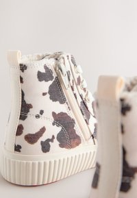 Kreminės spalvos aukštos drobinės sportbačiai su rudų ir juodų karvės raštais. Yra užtrauktukas, tekstūruotas guminis padas ir minkštas pamušalas.