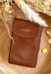 Brązowy skórzany portfel z lekkim połyskiem, z przednią kieszonką i złotym wytłoczonym logo. Otoczony monetami i suszonymi kwiatami na drewnianej powierzchni.