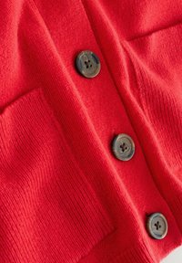 Red knitted cardigan featuring two large buttons and patch pockets. Texture is soft with ribbed detailing along the edges.