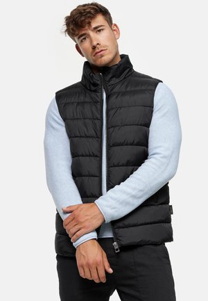 Zwart gewatteerd vest met een hoge kraag en een rits aan de voorkant, gedragen over een lichtblauw gestructureerd langemouwen shirt. Het model poseert met gekruiste armen.
