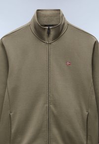 Chaqueta verde oliva con cremallera, hecha de tejido suave y con un cuello alto. Presenta un pequeño emblema de la bandera noruega en el pecho y bolsillos laterales.