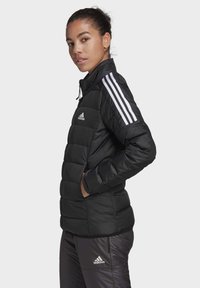 Chaqueta acolchada negra con diseño de cuadrados, cierre de cremallera, cuello alto y acentos en blanco con tres rayas en los hombros. Incluye el logo de Adidas.