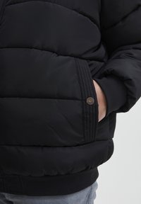 Sort polstret jakke med quiltet design, fremhævet af ribbet manchet og trykknapdetalje på sidelommen. Glat tekstur.
