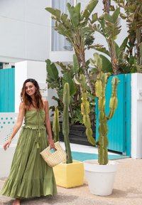 Green tiered maxi dress with a drawstring, carrying a cream woven bag. Background features tropical plants and a blue wooden gate.