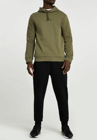 Felpa verde oliva con cappuccio e cordino, logo ricamato, abbinata a pantaloni jogger neri e sneaker nere. I materiali sembrano morbidi e strutturati.