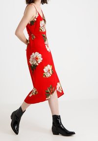 Röd blommig midiklänning med tunna band och en avslappnad passform, med vita och gröna blomsterdesigner. Bärs med svarta ankellägenheter.