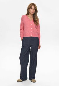 Rozā vilnas kardigāns ar V-veida kakla izgriezumu un pogu aizdari, apvienots ar tumši ziliem platām kājām džinsiem un bēšiem slip-on apaviem.