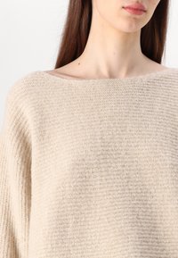 Šviesiai rudas megztinis su plačia, plačia iškirpte ir tekstūruotu paviršiumi. Audinys atrodo minkštas ir jaukus, su laisvais rankovėmis.