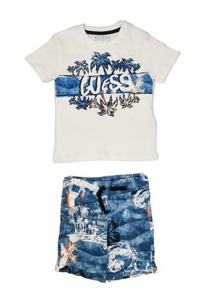 T-shirt bianco in cotone con grafica di palma blu e testo "GUESS", abbinato a pantaloni corti blu con stampa tropicale e vita con cordoncino.