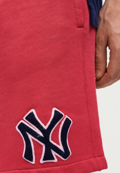 Κοντινό πλάνο χεριού ατόμου δίπλα σε κόκκινα σορτς με ραμμένο μπλε-σκούρο και λευκό λογότυπο "NY" κοντά στο τελείωμα.