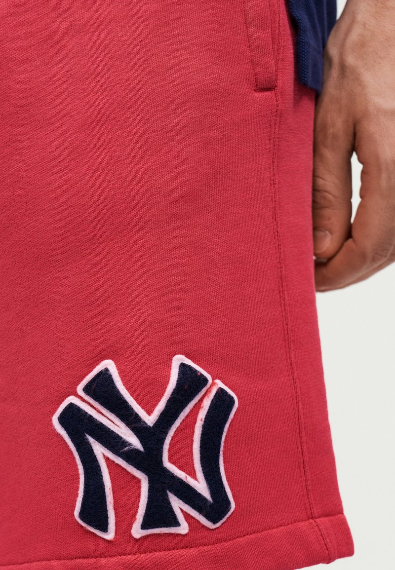 Close-up van de hand van een persoon naast rode korte broek met een gestikte marineblauwe en witte "NY"-logo vlak bij de zoom.