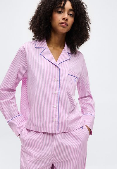 Femme aux cheveux bouclés portant un pyjama rayé verticalement rose et blanc avec un liseré bleu, debout les mains dans les poches devant un fond uni.