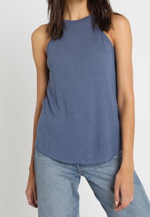 Blauwe mouwloze top van zacht materiaal, met een hoge halslijn en een gebogen zoom, gecombineerd met lichte blauwe spijkerbroek.