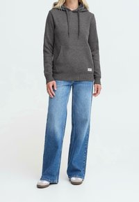 Personne portant un sweat à capuche gris foncé, un jean large bleu clair et des baskets blanches, debout contre un fond clair uni.
