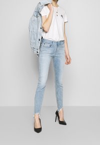 Persona que lleva vaqueros ajustados de color azul claro con dobladillos deshilachados, una camiseta blanca, tacones altos negros y sostiene una chaqueta de mezclilla sobre el hombro.