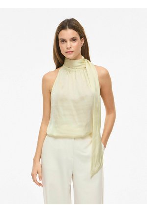 Kvinde iført en bleg gul ærmeløs haltertop med et langt sideskørt kombineret med højtaljede hvide bukser mod en hvid baggrund.