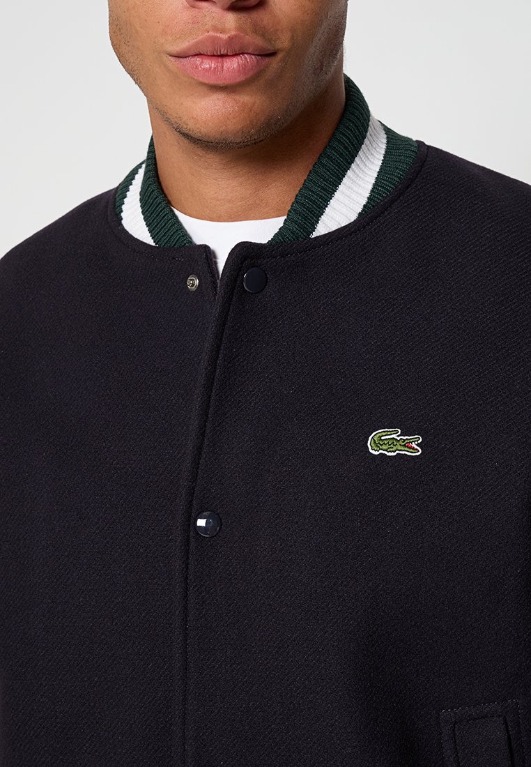Veste de varsity noire avec un col rayé vert et blanc, fermeture à boutons-pression, et un logo de crocodile brodé sur la poitrine gauche.