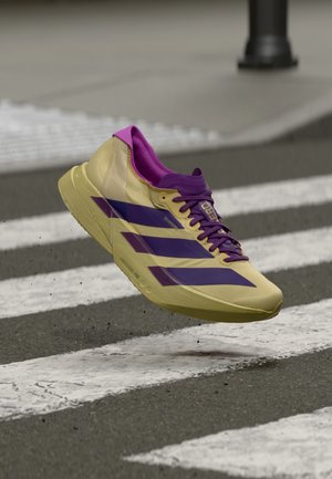 Leichter Laufschuh in Gelb mit violetten Akzenten. Verfügt über ein schlankes Design, eine strukturierte Oberfläche und ein auffälliges Drei-Streifen-Logo.