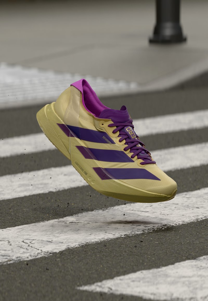 Chaussure de course légère jaune avec des accents violets. Présente un design élégant, une surface texturée et un logo à trois bandes bien visible.