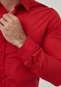 Chemise rouge à boutons dans un style ajusté, fabriquée en tissu lisse avec un col structuré et des manches longues, dotée de poignets boutonnés.