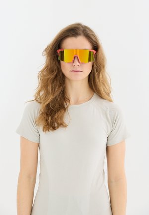 Femme aux longs cheveux ondulés porte des lunettes de sport réfléchissantes orange et un t-shirt athlétique beige clair à manches courtes, regardant vers l'avant.