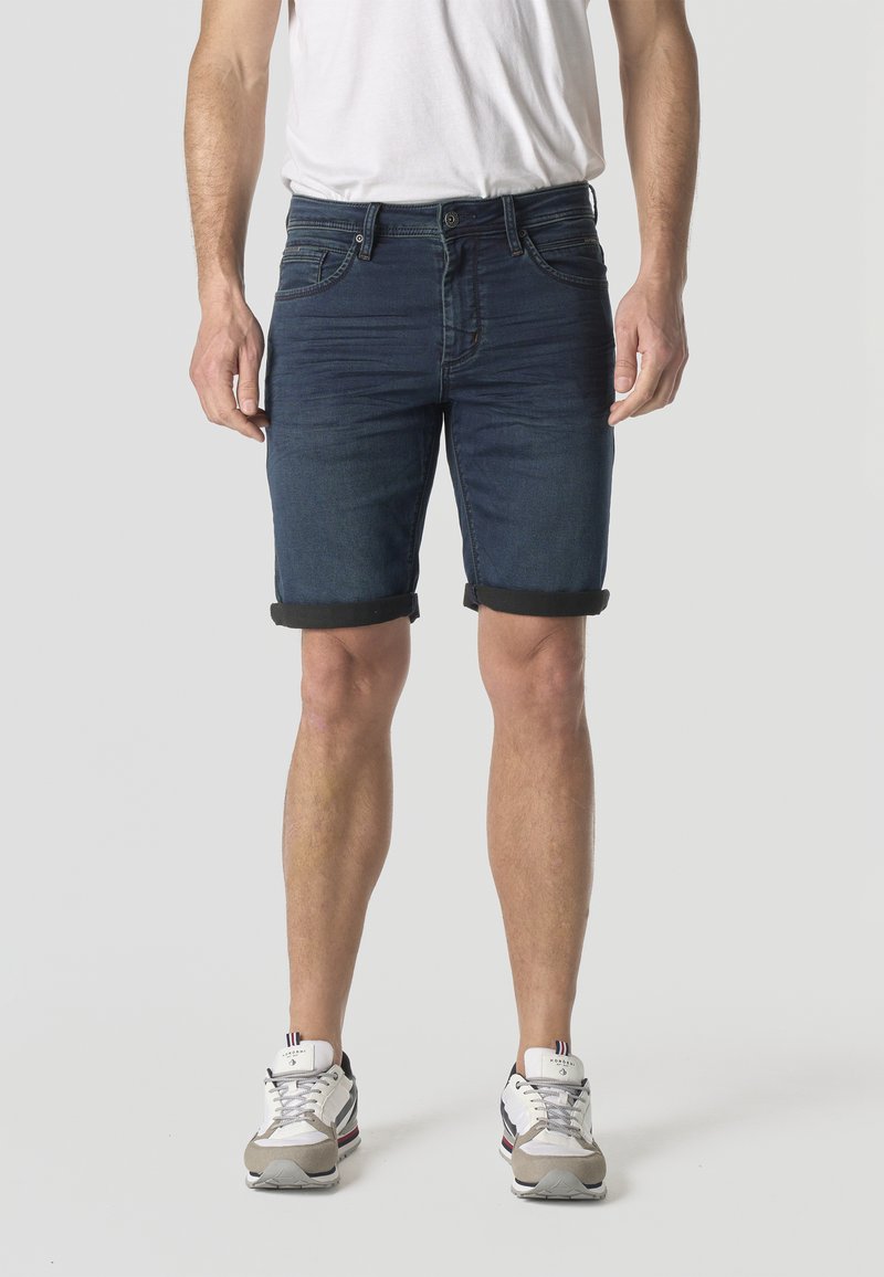 Férfi sötétkék felhajtott farmer rövidnadrágban, fehér pólóban és fehér tornacipőben, világosszürke háttér előtt állva.