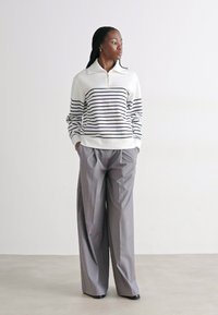 Pullover bianco con righe navy, colletto con zip; abbinato a pantaloni larghi grigi chiari. Tessuto morbido, vestibilità rilassata.
