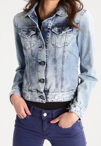 Ljust blå denimjacka med blekta partier, knäppning fram och bröstfickor. Har en fransad nederkant och textilstruktur. Matchas med mörkpurpurfärgad byxor.