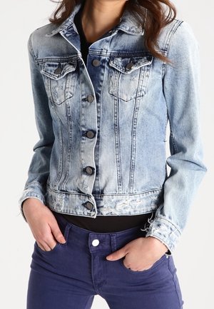 Chaqueta de mezclilla azul clara con áreas desteñidas, cierre de botones y bolsillos en el pecho. Presenta un dobladillo deshilachado y un tejido texturizado. Combinada con pantalones morados oscuros.