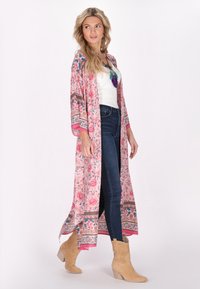 Kimono floreale rosa lungo fino ai piedi con motivi intricati, maniche lunghe e bordi in contrasto, abbinato a jeans scuri e stivaletti color nocciola.