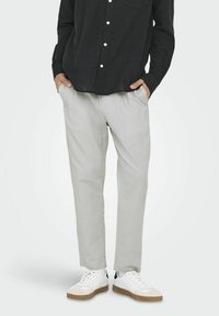 Pantalones gris claro hechos de una tela suave y texturizada, con bolsillos laterales y un corte relajado. Combinados con zapatillas blancas y una camiseta negra.