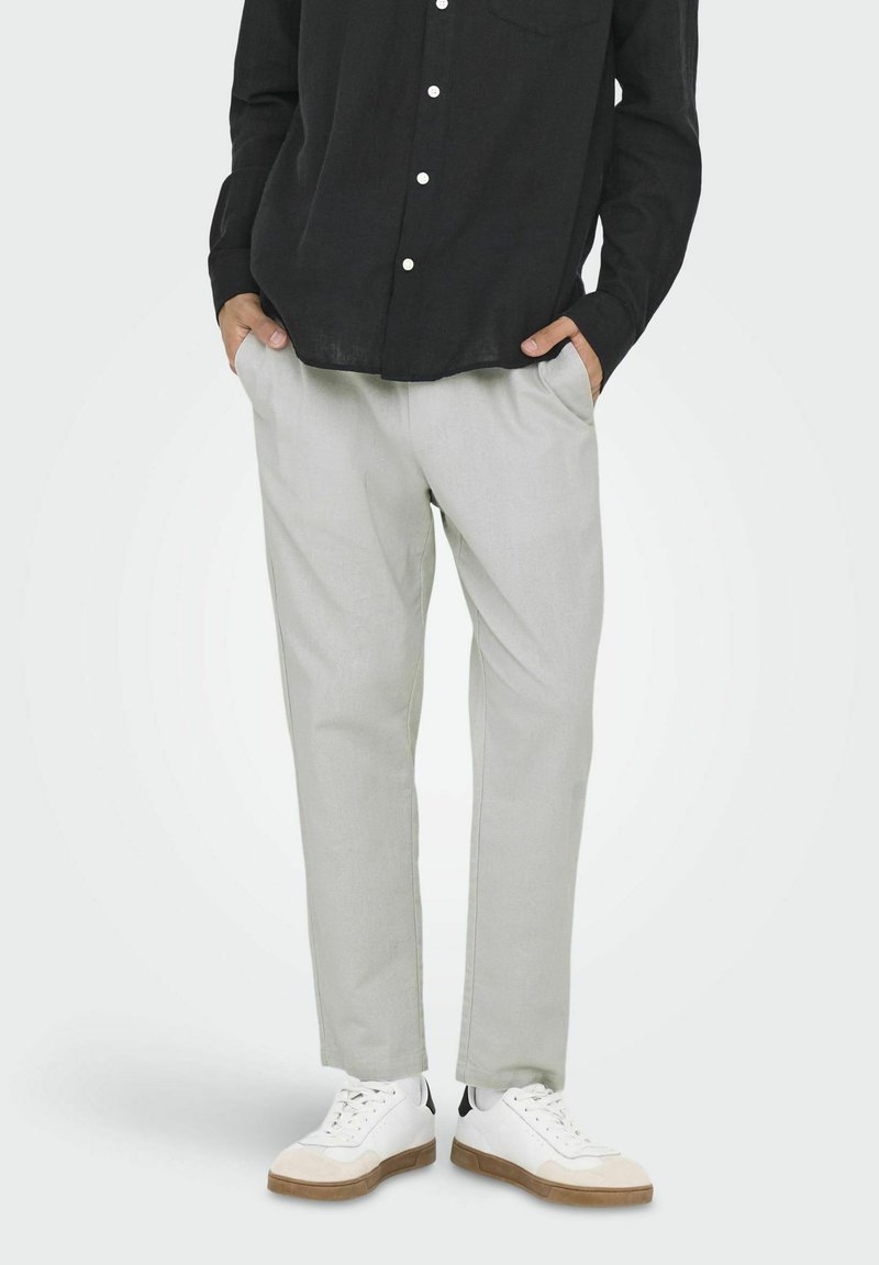 Pantalones gris claro hechos de una tela suave y texturizada, con bolsillos laterales y un corte relajado. Combinados con zapatillas blancas y una camiseta negra.