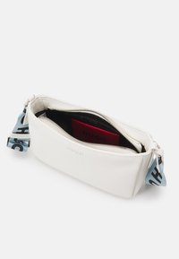 Bílá obdélníková crossbody taška se stříbrným zipem, černým vnitřkem, červenou nálepkou a modrým látkovým popruhem s černým textem.