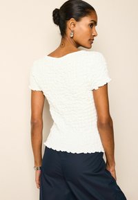 Haut blanc texturé avec des manches courtes et des bords festonnés, ajusté à la taille. Porté avec un pantalon sombre et ample.