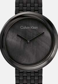 Črni metalni časovni izdelki z gladkim, temno sivo številčnico, minimalističnim dizajnom in blagovno znamko Calvin Klein. Ponuja povezano črno zapestnico in preproste kazalce ure.