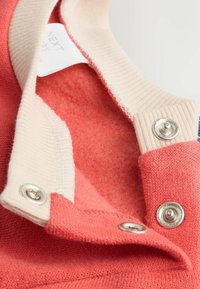 Koralsweater med krave i creme farve og sølvknapper; blød indvendig foring med synlige syninger. Tagget læser "NEXT." Detaljerede sømme accenter.