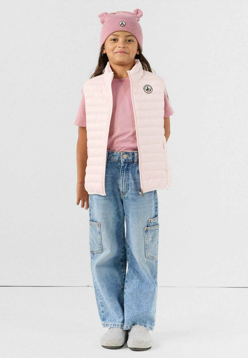 Lichtroze gewatteerd vest over een roze shirt met korte mouwen, gecombineerd met wijde blauw denim broek en een roze gebreide muts met berenoren.