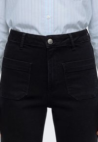 Taille et parties supérieures des jambes portant un pantalon en jean noir avec de grandes poches avant et une chemise bleue rayée rentrée, vue centrée.