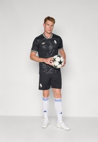 Sort fodboldtrøje med korte ærmer, har grafisk print, hvide accenter, parret med sorte shorts, hvide sokker med blå striber, holder en fodbold.