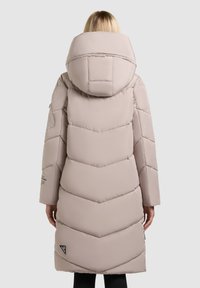 Kvinde iført en lang, lys beige polstret vinterjakke med en stor hætte, set fra ryggen mod en neutral baggrund.