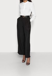 Bílá blůza s vlnkovým výstřihem a dlouhými rukávy, kombinovaná s černými kalhotami s vysokým pasem a širokými nohavicemi, a černými sandály na podpatku.