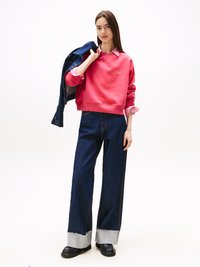 Felpa rosa con polsini a coste, indossata sopra una camicia a righe con colletto. Jeans a gamba larga in denim scuro con orlo grezzo, abbinati a mocassini neri.