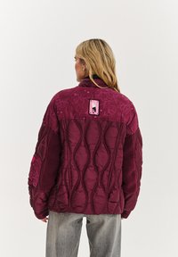 Bordový prešívaný kabát s textúrovanými panelmi, kvetinovým vzorom a ružovým logom. Má vysoký golier a voľný strih.